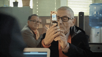 Movie gif. Sergio Chamy in The Mole Agent, holds a flip phone, squinting at it as he turns it over in his hands.