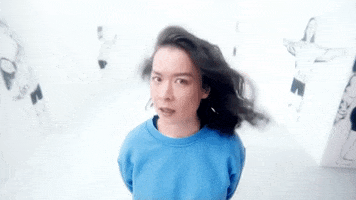 Music video gif. Wearing a bright blue sweater, Mitski stands in a blindingly white room that has faded black and white portraits decorating the walls. She shakes her head at us while her hair blows in the wind.
