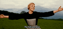 Movie gif. Julie Andrews as Maria in The Sound of Music sings in a hilly field with a vista of mountains, holding her arms out joyfully as she sings, "The hills are alive."