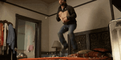 TV gif. Charlie Day as Charlie in It’s Always Sunny in Philadelphia jumps on his bed while eating popcorn, recreating a scene from Home Alone.