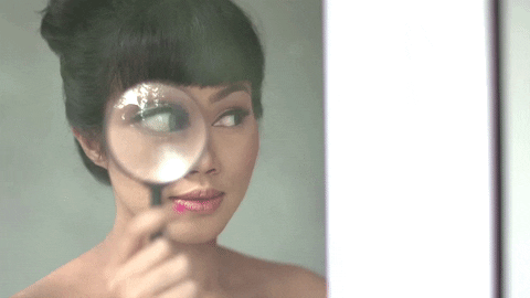 An animated gif video of a woman holding a magnifying glass over one eye, so that it's larger. She holds her head still and just moves her eyes side-to-side, like she's looking for something shifty.