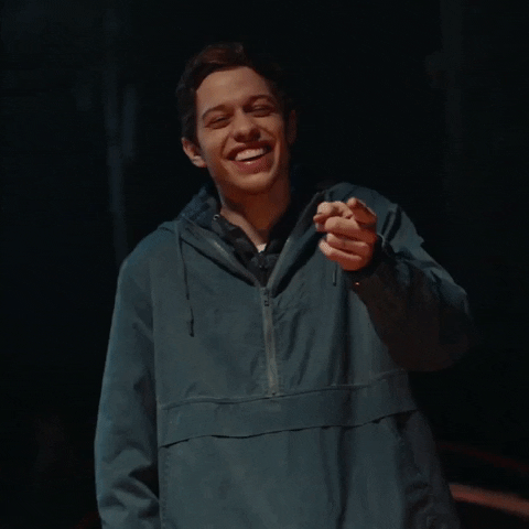 SNL gif. Comedian Pete Davidson laughs next to a campfire at night. 