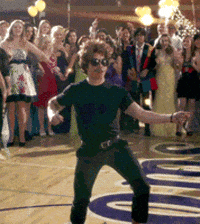 Video gif. A man in a black T-shirt and pants swinging his hips on a dance floor in a high school gym, surrounded by students in formal wear.
