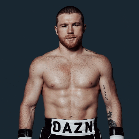 Sports gif. Canelo Álvarez, a Mexican Boxer, is shirtless and wearing his boxing shorts and gloves. He stands in front of us and gives us a smile and a wink.