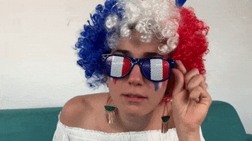 Video gif. A woman wearing a curly wig and sunglasses, both striped like the French flag, tilts her shades and leans in for a closer look at us.