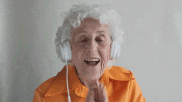 Video gif. An elderly woman wears headphones and grins cheerfully as she slowly claps. 