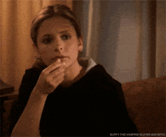TV gif. Sarah Michelle Gellar as Buffy sits on a couch watching something and absentmindedly eating popcorn.