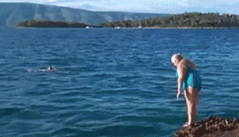 Video gif. Woman stands at the edge of a stony bank making a half-hearted attempt at a dive, bellyflopping into the water.