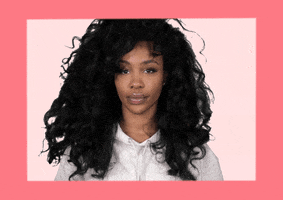 Celebrity gif. Singer Sza looks at us and places her hand on her chest. She says with almost a flirtatious gaze, “Who me?”