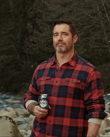 Ad gif. Man with a beard and a plaid button up shirt stands next to a rocky river in the wilderness. He looks at us with a smug smile, holding a Busch beer in one hand, giving a big thumbs up with the other.