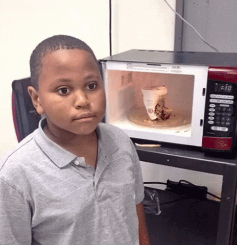 Video gif. Zoom in on a boy standing in front of a microwave with a blank, empty stare. In the open microwave, is a burnt up instant ramen cup. Text, “What is Life?”