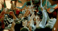 Video gif. Partygoers bounce around with martini glasses lifted in the air as a woman haphazardly pours champagne from a jug.