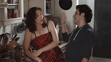 Video gif. Woman leans against a kitchen counter, looking annoyed as a man holds his hands up for a high five from her. He slaps her arm and then holds his hand up, waiting for the high five.