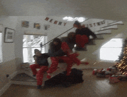 Video gif. Teenagers wearing Christmas pajamas sit on the edge of a staircase without a railing as two topple off in a room decorated for the holidays.