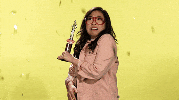 Celebrity gif. Awkwafina holds a trophy and shudders as she sees confetti flying around her. Then, she embraces it and smiles and waves at us while playing with the floating confetti.