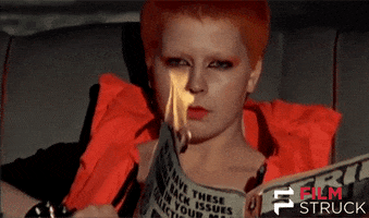 Video gif. A person with short orange hair and dramatic orange makeup holds a magazine with a corner lit on fire. A smile gradually emerges on their face as they look past the flame at us.