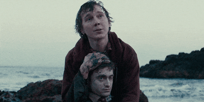 Video gif. Patrick Dano as Hank and Daniel Radcliffe as Manny in Swiss Army Man sit on the beach. Hank holds Manny's head in his arms as he speaks sincerely to someone off screen. 