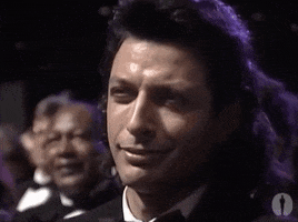 TV gif. a long-haired Jeff Goldblum at the 1989 Oscars smiles and gives a little nod with a confident, dashing look on his face. 