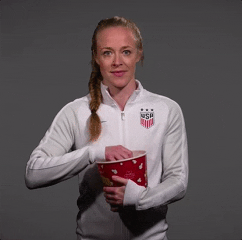 Sports gif. Becky Sauerbrunn holds a big tub of popcorn and stuffs her mouth with popcorn while staring at us.