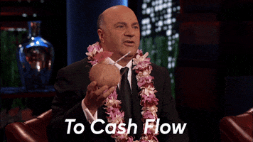 Reality TV gif. Kevin O'Leary on Shark Tank wearing a lei and holding a drink in a coconut. He toasts us and says, "To cash flow!"