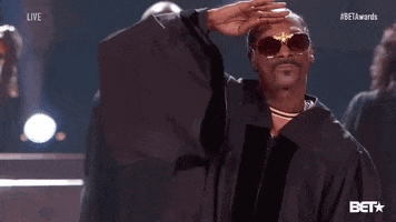 Celebrity gif. Snoop Dog wears a black choir robe and dark, ornate sunglasses at he salutes to us. Women in choir robes sing and clap behind him.