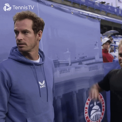Sports gif. Andy Murray and another are on the sidelines in street clothes. Andy looks over his shoulder, flicking his tongue side to side in a playful way as the other gives a thumbs down, looking in the same direction. 