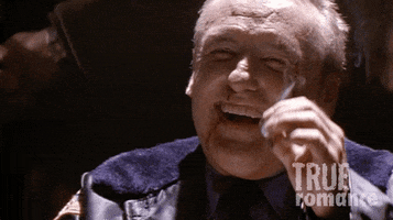 Movie gif. Dennis Hopper as Clifford in True Romance. He's beaten up and sitting on a chair, cackling. He raises his cigarette in toast to the person and takes a puff.