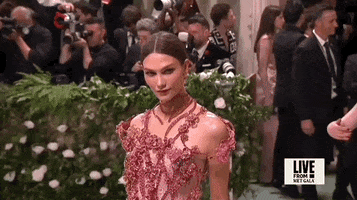 Met Gala 2024 gif. Close up of the top of Karlie Kloss wearing a jeweled pink floral Swarovski gown with ropes of fabric weaving an intricate design across her torso and around her neck. 