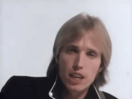 Music video gif. Tom Petty sings as he looks at the camera, playing a guitar out of frame. Text, "Yeah the waiting is the hardest part," with emphasis on the word hardest. 