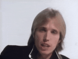 Music video gif. Tom Petty sings as he looks at the camera, playing a guitar out of frame. Text, "Yeah the waiting is the hardest part," with emphasis on the word hardest. 