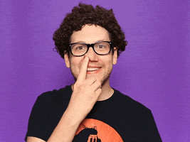Video gif. Nerdy man twists a finger in his nose as he smiles blankly at us.