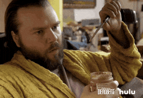 TV gif. Tyler Labine as Kevin Pacalioglu on Deadbeat sits in a chair in a robe. He holds a jar of peanut butter and a butter knife. He nods and winks as he chews on peanut butter. 