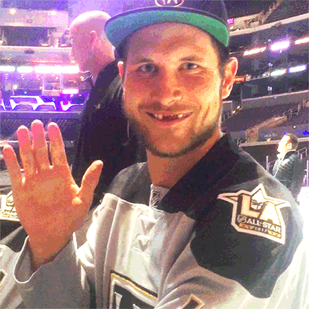 Sports gif. Kyle Clifford wearing his LA Kings hockey jersey and a ball cap, sits in an arena, smiling and waving at us with three missing front teeth.
