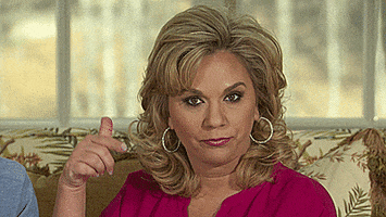 Reality TV gif. Julie Chrisley on Chrisley Knows Best is sitting on a couch and she rolls a finger at her temple, making the crazy symbol, while staring at us.