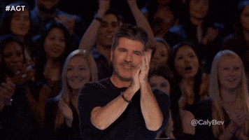 Celebrity gif. Simon Cowell stands up after a performance and enthusiastically applauds and flashes two thumbs up.