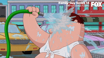Family Guy gif. Peter Griffin stands in a lot of cars wearing a woman's bathing suit top while he smiles and turns his head back and forth, pouring water from a hose on his face. 