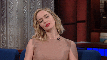Celebrity gif. Emily Blunt as a guest on the Late Show with Stephen Colbert raises her hand in a peace sign, her head cocked to the side as she purses her lips halfheartedly.