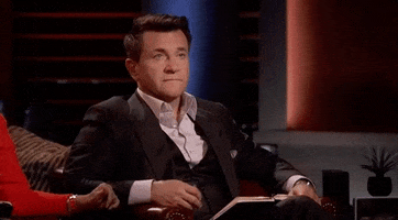 Reality TV gif. Robert Herjavec on Shark Tank is sitting in his chair and looks unpleased. He has a stoney face as he raises his hands palms up, shrugging and unbothered.