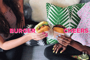 Video gif. Two women sit next to each other on a sofa and move their burgers towards each other like they’re clinking glasses together. Text, “Burger cheers.”