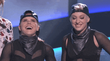 Video gif. Two women in identical outfits with black backwards caps, leather scarves, and tops appear completely flattered with humongous smiles spread across their faces, the rightmost one clutching her hand to her chest.
