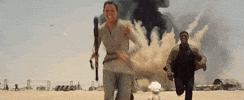Star Wars gif. Daisy Ridley as Rey and John Boyega as Finn in Star Wars: The Force Awakens run away from a massive explosion in the desert.