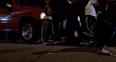 Movie gif. Large group of men fight in a parking lot in front of a red pickup truck. A black fedora hat falls off one of man and rolls around in the foreground.