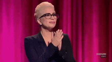 Reality TV gif. Lady Gaga on Rupaul’s Drag Race wears a pair of simple glasses and a black suit jacket. She puts her hands up to her mouth and kisses them, then takes her hands away from her mouth to send the kiss off to someone. She has a big, proud smile on her face. 