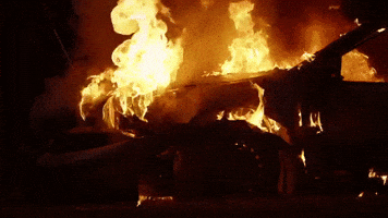 Video gif. Dilapidated car is on fire at night.