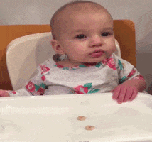 Video gif. Baby in a high chair slams its hand on the eating tray alongside a stray Cheerio, propelling the Cheerio directly into its mouth and catching it perfectly in its lips.