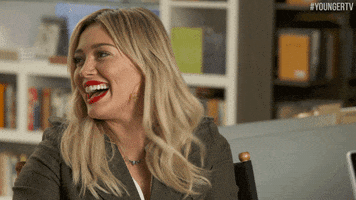 TV gif. Hilary Duff as Kelsey Peters from Younger, seated, leans forward and laughs uproariously.