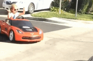 Video gif. Preschool age girl in sunglasses holds up both hands with 2 fingers up in a rock on gesture. She rides in a red toy sports car that turns and slides out into the street. 