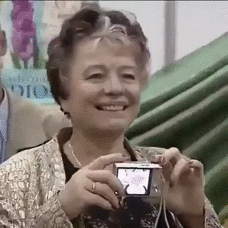 Video gif. An older woman in a shiny jacket looks at something right of frame, and cheerfully holds up a small digital camera to take a picture, but the camera's view screen is facing away from her. The flash lights up as she takes a picture of herself.