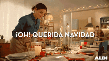 Video gif. Woman with an apron on leans over a fully decorated table in a dining room. A man is in the kitchen, at the counter cooking something. Text, “Oh! Querida Navidad.”