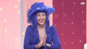 Video gif. A woman wearing a matching indigo frilly hat, wig, eyeliner, and dress grins and glances to the side while clapping her hands rapidly. 
